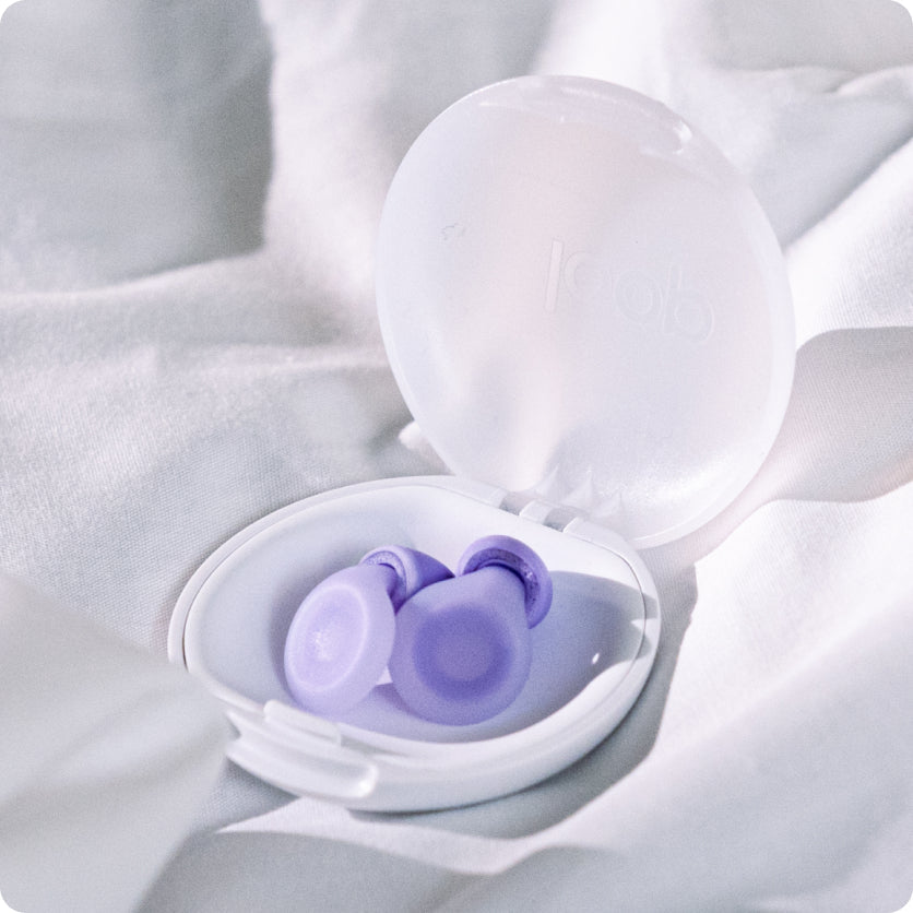 Lilac Loop Dream earplugs in their open white case on bedding